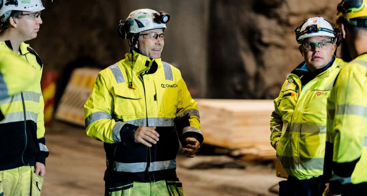 A group of OneCo workers are seen meeting together on the job with helmets and uniforms on.