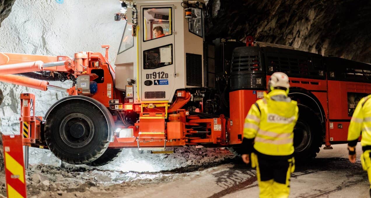 A drilling machine is pictured digging out the side of a tunnel. The back of two workers are visible in the foreground.