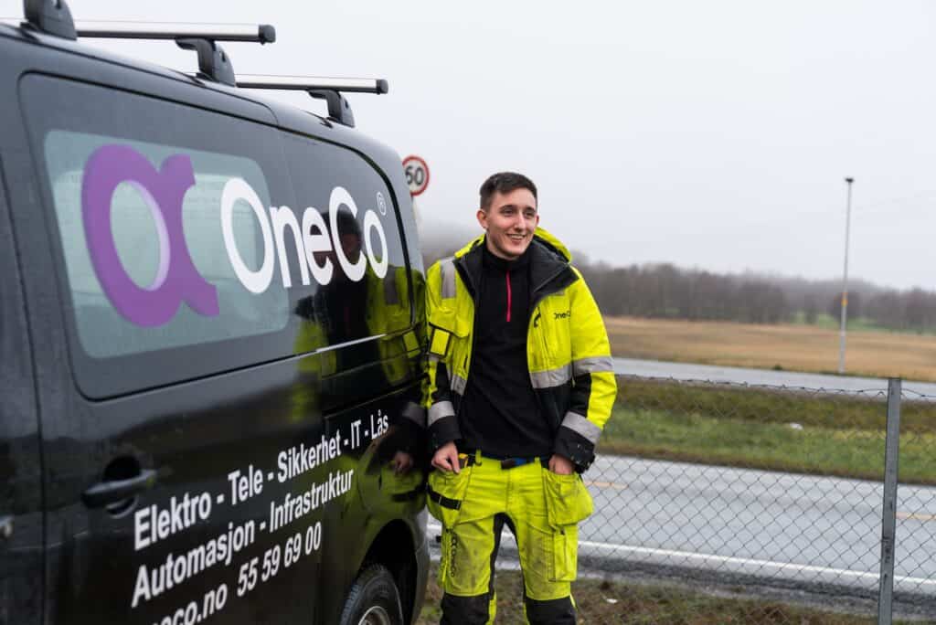 OneCo lærlinger leaning against his work car with hands in pockets smiling into the distance